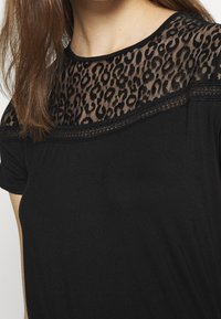 Černé tričko s průhlednou krajkovou vložkou, která má design leopardího vzoru na výstřihu, strukturovaná tkanina a uvolněný střih.