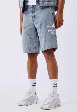 Man draagt lichtblauwe denimshorts met wit logo op zijn linkerbeen, witte sokken met zwart logo en witte sneakers, staand op een witte achtergrond.