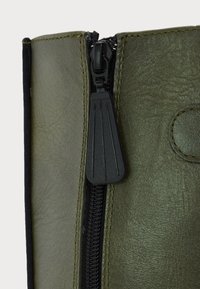 Gros plan sur une tige de botte en cuir vert olive avec une fermeture éclair noire et un grand tirette de fermeture éclair.