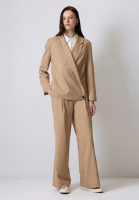 Peščena blazer s ovitenim dizajnom in eno gumbom, v kombinaciji z usklajenimi širokimi hlačami, ki imajo subtilno teksturo in kroj po meri.