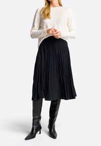 Suéter blanco texturizado de mangas largas combinado con una falda midi negra con plisado. Botas negras de tacón alto y puntera afilada hasta la rodilla completan el look.