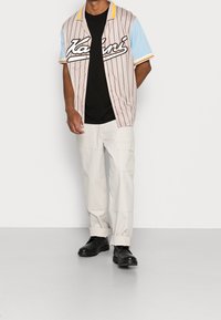 Uomo che indossa una camicia beige a righe con maniche blu, una maglietta nera, pantaloni cargo bianchi e scarpe nere, in piedi contro un muro bianco.