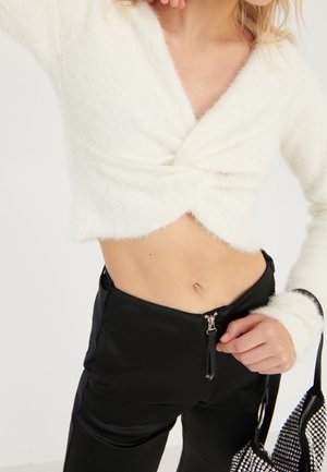 Femme portant un pull court blanc duveteux avec un devant torsadé et un pantalon noir taille haute, tenant un sac à main noir clouté.