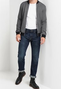Chaqueta bomber gris con puños y cuello acanalados negros, camiseta blanca, vaqueros oscuros con puños remangados, botas negras.