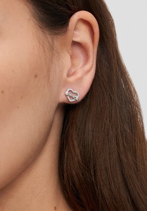 Nahaufnahme eines Frauenohrs mit einem silbernen steckförmigen Ohrring in Herzform, der Kristalle enthält und eine diagonale Leiste über das Herz verläuft.