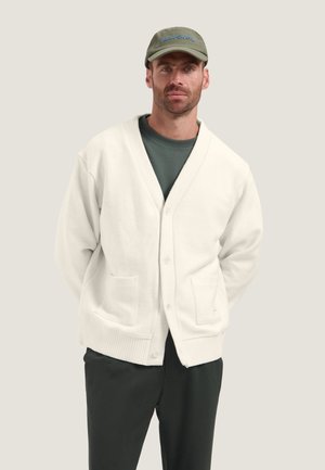 Mand med skæg iført grøn kasket, mørk skjorte, hvid knaplukket cardigan med lommer og mørke bukser stående mod en ensfarvet lys baggrund.