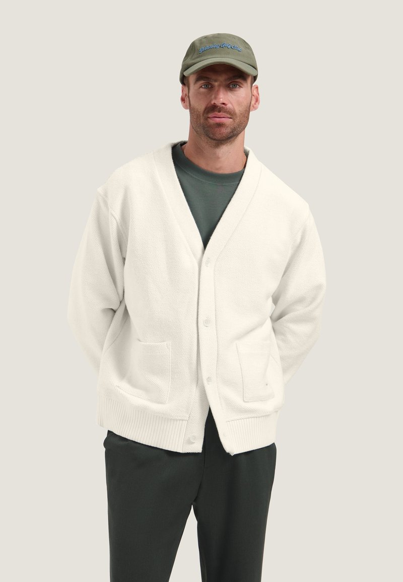 Homme barbu portant une casquette verte, une chemise sombre, un cardigan blanc boutonné avec des poches, et un pantalon sombre, debout devant un fond clair uni.