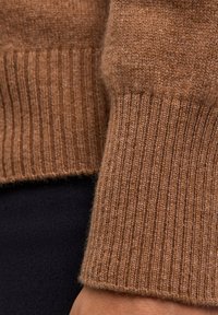 Brauner Strickpullover mit Details zu gerippten Bündchen und einer weichen Textur, kombiniert mit einem schwarzen Kleidungsstück darunter für den Kontrast.