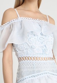 Světle modrý top bez ramínek s květinovým krajkovým designem, volánkovými rukávy a otevřenými detaily. Texturovaná látka s jemnými vzory.