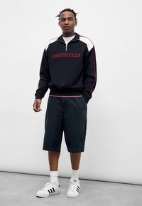 Sudadera negra con cremallera y texto rojo "LETS FRANS", combinada con pantalones cortos oscuros de rayas y zapatillas blancas Adidas con rayas negras.
