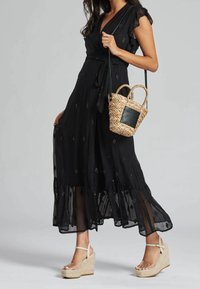 Schwarzes Wickelkleid mit Rüschensämlen, das zarte Verzierungen aufweist. Kombiniert mit einer geflochtenen Tasche und beigen Keilsandalen.