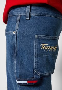 Pantaloni cargo in denim con lavaggio blu, dotati di tasca laterale, logo "Tommy Jeans" in rilievo color oro e un accento in tessuto multicolore sulla tasca.