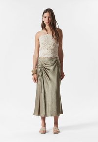 Beige strukturierter Crop-Top mit wirbelnden Mustern und einem olivgrünen asymmetrischen Rock mitraffbarer Detail. Flache Sandalen runden den Look ab.