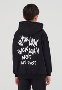 Felpa nera con cappuccio, tasca a marsupio frontale, vestibilità oversize e testo grafico bianco sul retro: "NON GUARDARE MAI INDIETRO ANCORA NON VELOCE VELOCE."