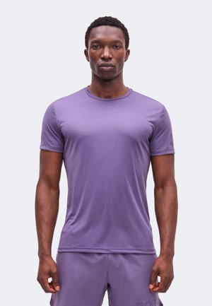 Muškarac stoji okrenut prema naprijed, nosi usku ljubičastu košulju kratkih rukava i odgovarajuće ljubičaste kratke hlače na jednobojnoj pozadini.