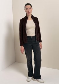 Femme debout contre un mur beige, portant une veste zippée marron foncé, un haut beige, un pantalon noir et des baskets blanches.