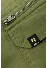 Tkanina w oliwkowym kolorze z zamkiem błyskawicznym oraz czarną metką z białym napisem. Teksturowana powierzchnia z widocznymi detalami szwów.