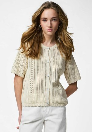 Młoda kobieta z długimi włosami, ubrana w kremowy, krótki swetrowy kardigan na guziki oraz białe spodnie, stojąca na jednolitym tle.