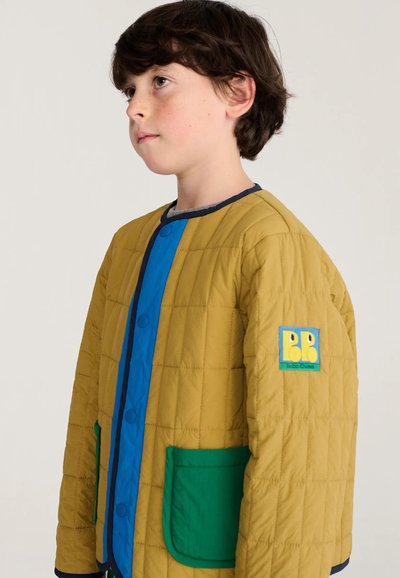 Enfant aux cheveux courts et foncés portant une veste matelassée moutarde avec des bordures bleues et des poches vertes, regardant sur le côté.