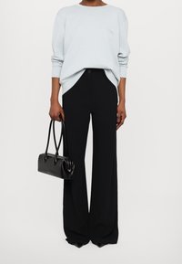 Kvinna som bär en ljusblå långärmad topp, svarta vida byxor, och håller en svart handväska med dubbla handtag.