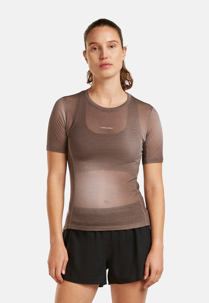 Vrouw die recht vooruit staat, gekleed in een strakke, bruine, gestructureerde top met korte mouwen en losse zwarte shorts, tegen een witte achtergrond.