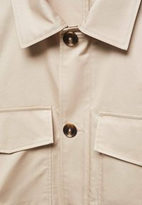 Beżowa koszula z klasycznym kołnierzykiem, dwoma kieszeniami na piersi i brązowymi guzikami, wykonana z gładkiej tkaniny z czystym wykończeniem szwów.