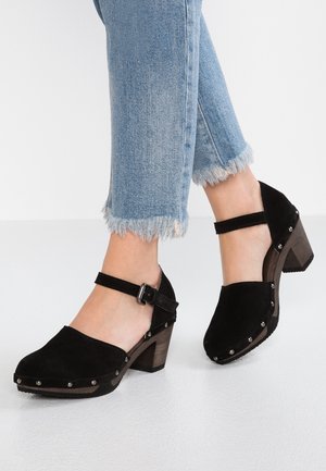 Czarne zamszowe buty na obcasie z paskiem wokół kostki, okrągłym noskiem i drewnianym słupkowym obcasem. Ozdobione dekoracyjnymi srebrnymi ćwiekami wzdłuż podeszwy.