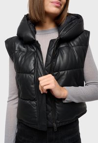 Gilet in pelle nera con un design trapuntato, cappuccio over-size e chiusura frontale con zip. Indossato sopra una maglietta grigia chiara a maniche lunghe con coste.