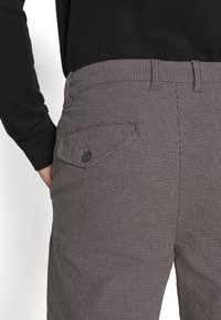 Man i grå rutiga byxor med en knappad bakficka och en svart långärmad skjorta, med ena handen delvis i sidfickan.