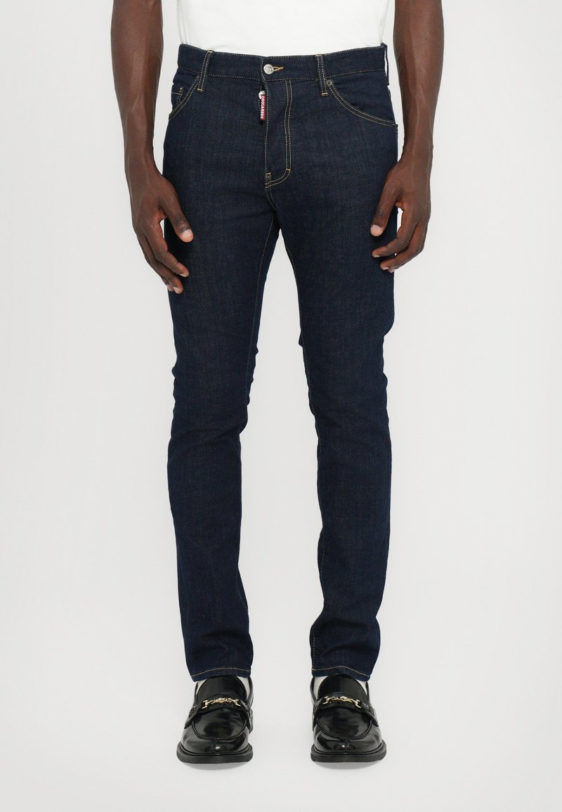 Jean en denim bleu foncé, coupe slim, avec détails de couture, passants de ceinture et une petite étiquette logo à l'avant. Matériau en coton avec une légère élasticité.