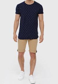 Camiseta de manga corta azul marino con lunares blancos, combinada con pantalones cortos caqui arremangados y zapatillas blancas. Diseño casual, material de algodón.