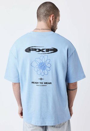 Man met kort haar en een ketting, draagt een lichtblauw oversized T-shirt met zwart grafisch ontwerp en de tekst "PXG" en "Ready to Wear New Classics".