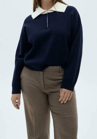 Personne portant un haut bleu marine à manches longues avec un col blanc et une fermeture éclair, associé à un pantalon marron à carreaux taille haute.