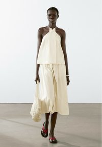 Lichtbeige mouwloze top met een halternek, gecombineerd met losse beige broeken. Het model draagt een geweven tas en heeft zwarte sandalen met rode banden aan.