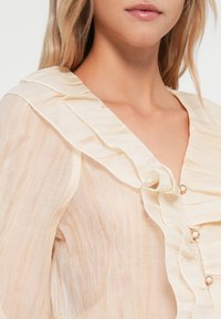 Gjennomsiktig, lys beige bluse med volangkant, subtile vertikale striper og dekorative gullfargede knapper langs fronten.