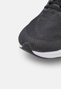 Gros plan de la pointe d’une chaussure de course noire et grise avec un tissu tricoté texturé et une semelle blanche sur fond blanc.