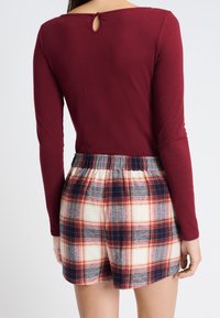 Top rojo de manga larga con un detalle en la parte posterior tipo ojal; combinado con pantalones cortos de cuadros en crema, azul marino y rojo, con una cintura elástica.