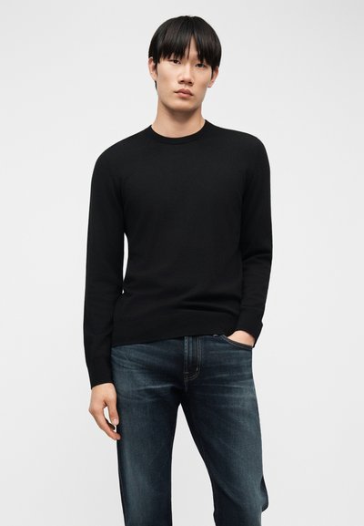 Νέος άνδρας με ίσια μαύρα μαλλιά φοράει μαύρο πουλόβερ μακρυμάνικο και σκούρο μπλε τζιν παντελόνι, στέκεται με το ένα χέρι στην τσέπη μπροστά σε απλό φόντο.