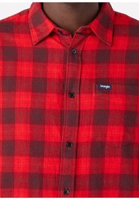 Chemise en flanelle rouge et noire avec un motif à carreaux, devant à boutons et poche. Comprend des boutons noirs et une petite étiquette Wrangler sur la poitrine.