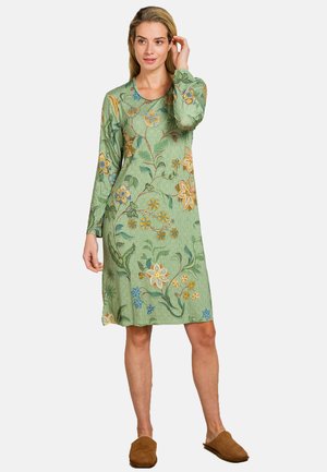 Vrouw in een groene lange mouw, jurk tot de knie met meerkleurige bloemenpatronen en bruine instappers, staand voor een witte achtergrond.