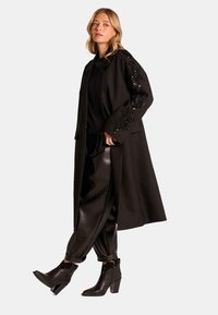 Abrigo largo negro con detalles de lentejuelas en las mangas, combinado con pantalones negros de tiro alto y botas de tobillo, todos mostrando un diseño elegante.