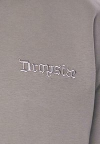 Grå sweatshirt med hvid broderet tekst "Dropsize," med en glat tekstur og tætsiddende design.