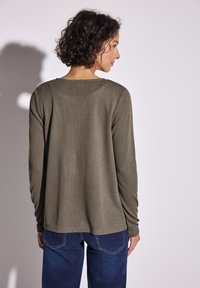 Frau mit kurzen lockigen Haaren, die ein olivgrünes Langarmshirt und dunkelblaue Jeans trägt, von hinten vor hellem Hintergrund.