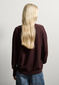 Pull en tricot marron à coupe décontractée, avec des poignets et un ourlet côtelés. Associé à un jean en denim, les cheveux tombent en vagues dans le dos.