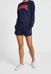 Námořnická mikina s červeným textem "HUMMEL", uvolněný střih, spárovaná s odpovídajícími námořnickými šortkami. Oblečení doplňují bílé tenisky.