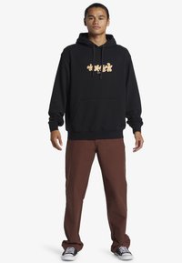 Sudadera negra con gráfico floral naranja, bolsillo canguro y cordones, combinada con pantalones marrones y zapatillas negras con suelas blancas.