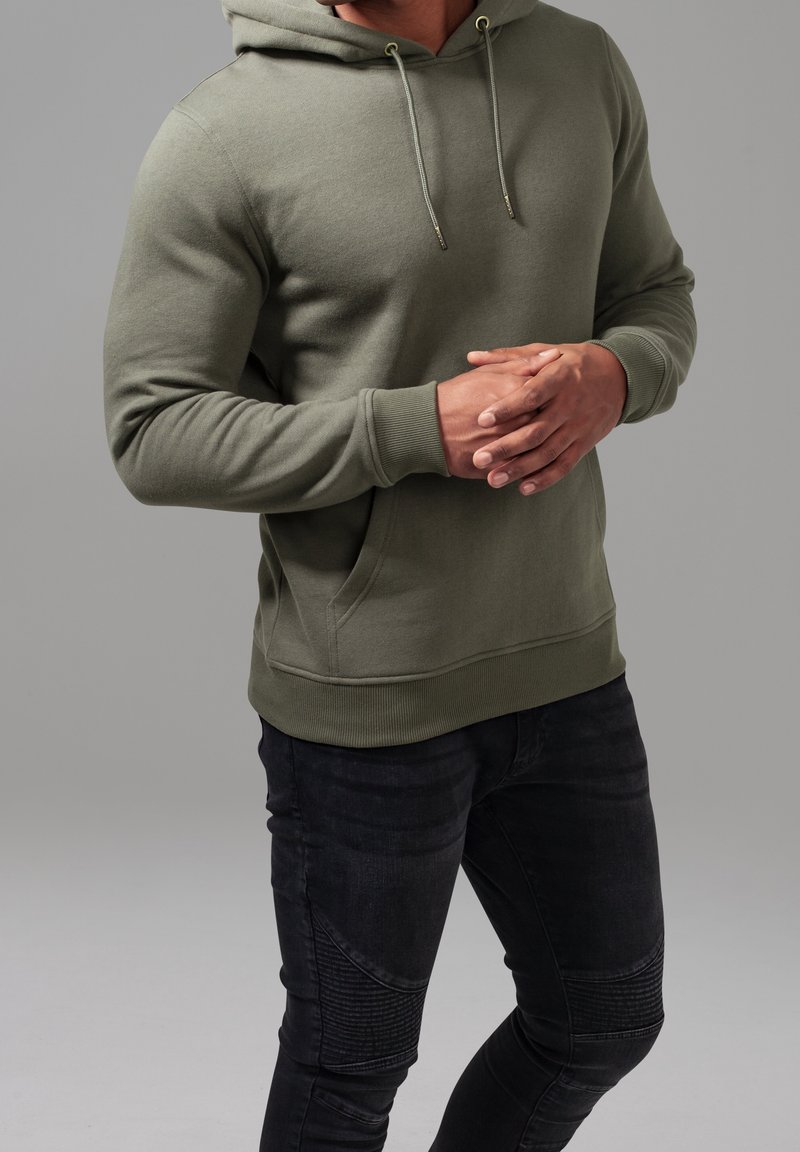 Homme portant un sweat à capuche vert olive et un jean noir texturé, debout les mains jointes devant un fond gris.