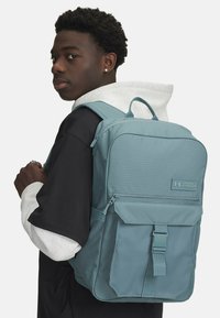 Teal fabric backpack with a rectangular design, front zipper pocket, and adjustable straps. Logo displayed on the front panel.