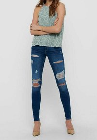 Débardeur floral vert clair associé à un jean bleu délavé moulant avec des déchirures et des accents effilochés, complété par des talons beiges.