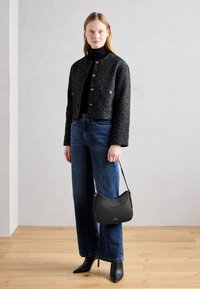 Nainen, joka on pukeutunut mustaan tekstuuriin takkiin, mustaan poolopaitaan, sinisiin farkkuihin ja mustiin kärjetöihin saappaisiin, ja pitää kädessään mustaa olkalaukkua.